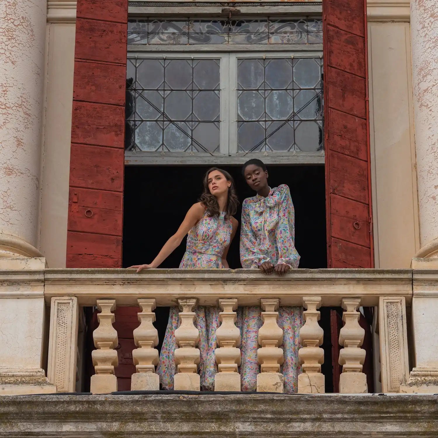 Two people wearing floral outfits standing on an ornate balcony.